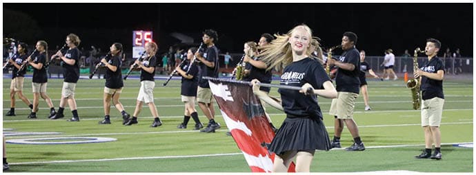 A person in a black outfit is twirling a flag on a sports field while a marching band in matching black shirts and khaki shorts plays instruments in the background. Descripci n generada con IA