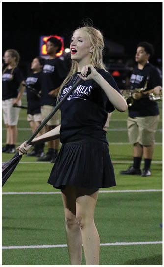 A person with long blonde hair wearing a black shirt and skirt is performing on a field with other people in the background playing musical instruments. Descripci n generada con IA