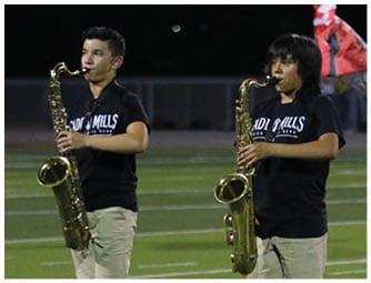 Three people wearing matching black shirts and khaki shorts are playing saxophones on a football field at night. Descripci n generada con IA
