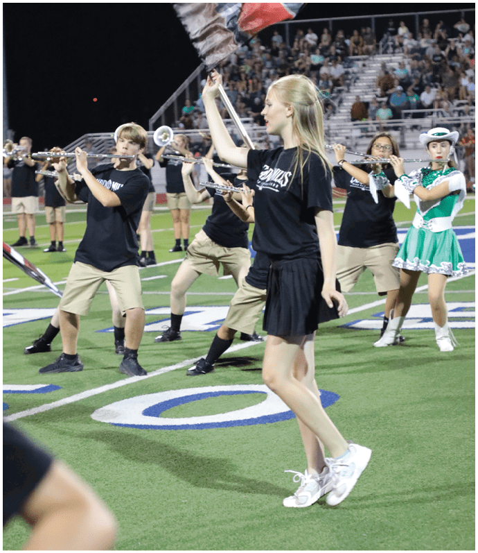 People wearing matching black shirts and khaki shorts are playing flutes and holding flags on a sports field at night with a crowd in the background. Descripci n generada con IA