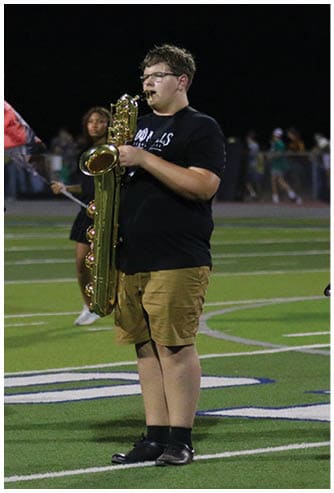 A person wearing a black t-shirt and khaki shorts is playing a saxophone on a sports field at night with another person holding a saxophone and a person waving a red flag in the background. Descripci n generada con IA