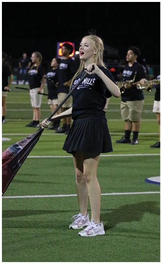 A person with long hair wearing a black shirt is holding a baton and appears to be singing or speaking passionately on a field with other people standing in the background. Descripci n generada con IA