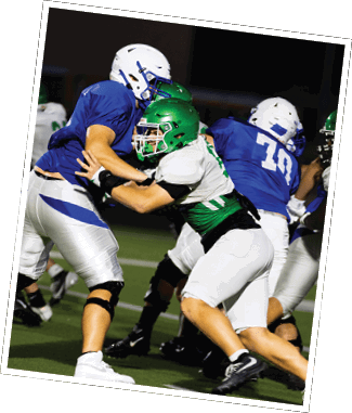 Two people in football uniforms are engaged in a blocking action on a field during a game at night. Descripci n generada con IA