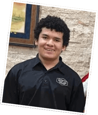 A person with curly hair wearing a black collared shirt with a logo stands smiling in front of a stone wall with a framed painting. Descripci n generada con IA