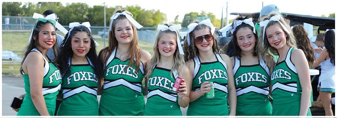 Seven people wearing green cheerleading uniforms with the word “FOXES" stand side by side outdoors. Descripci n generada con IA