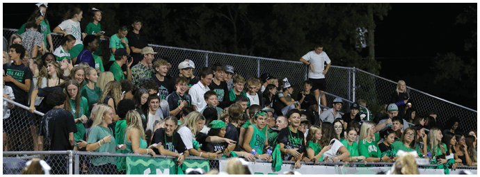 A large group of people wearing green and black clothing are gathered closely together in bleachers at an outdoor event during nighttime. Descripci n generada con IA