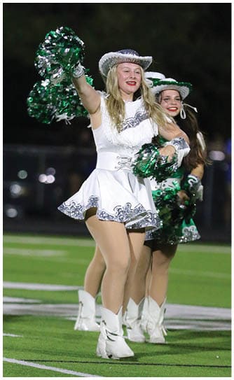 A person with long blonde hair is holding green and silver pom-poms, likely performing as a cheerleader against a dark background. Descripci n generada con IA