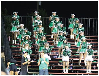 Adults in green and white dance uniforms perform a choreographed routine on a football field with a crowd and a drumline in the background. Descripci n generada con IA