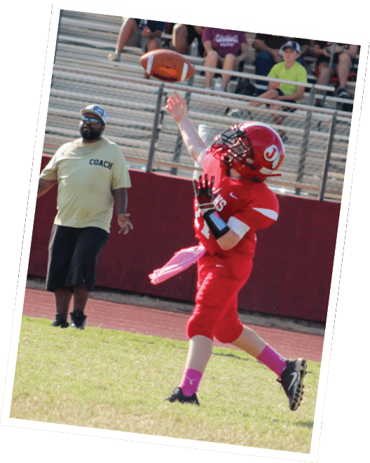 A person in red football gear is reaching up to catch a ball on a grassy field while another person wearing a coach shirt watches nearby. Descripci n generada con IA
