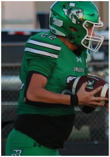 A person in green football gear is running on a field holding a football while three people stand in the background near a fence. Descripci n generada con IA