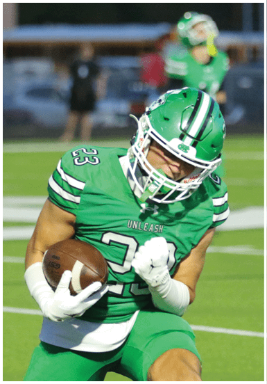 A person wearing a green football uniform and helmet is holding a football and appears to be running or preparing to make a play on a football field. Descripci n generada con IA
