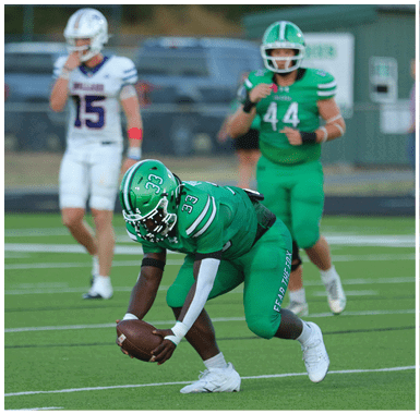 Two people in green football uniforms are on a field, with one person crouched and the other standing behind. Descripci n generada con IA