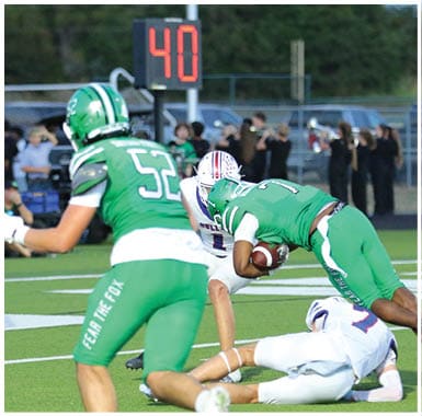 Two people in green football uniforms are actively playing on a football field with one holding the ball while another person in a white uniform is on the ground nearby. Descripci n generada con IA