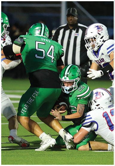 A group of people wearing football uniforms are engaged in a play on the field with one person in green holding the ball while others in white and green attempt to tackle or block. Descripci n generada con IA