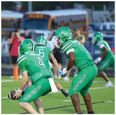 A person in a green football uniform with the number 4 is holding a football on a field with school buses and a person taking a photo in the background. Descripci n generada con IA