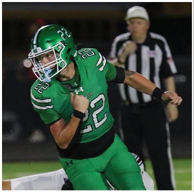 A person wearing a green football uniform and helmet is running on a field with a referee in a black and white striped shirt standing in the background. Descripci n generada con IA