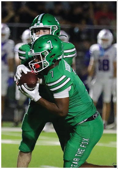 A person in a green football uniform with the number 7 is holding a football while another person in a similar uniform is behind them on a football field with blurred players in white uniforms in the background. Descripci n generada con IA