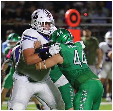 Two people in football uniforms are engaged in a play on a field with one person in green tackling or blocking the other in white. Descripci n generada con IA