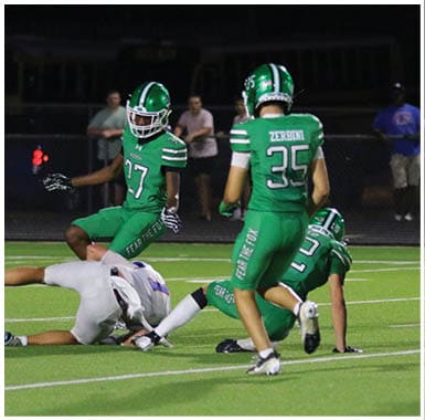 Four people in football uniforms are on a field with three in green uniforms and one in white, with two green-uniformed players standing and two on the ground during a game. Descripci n generada con IA