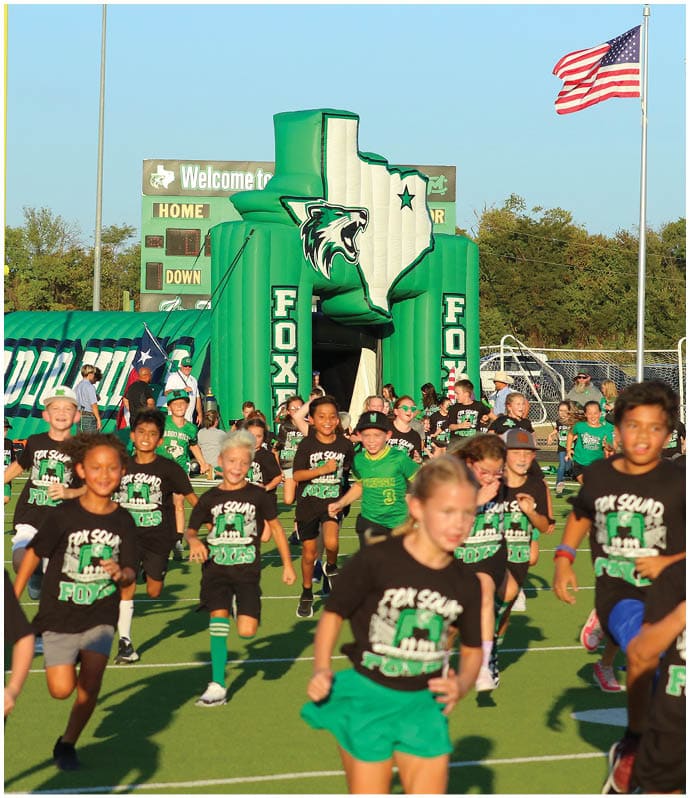 Children wearing matching green shirts are gathered near a large green inflatable structure shaped like the state of Texas with a wildcat logo and the word “FOX" on its sides, with an American flag visible in the background. Descripci n generada con IA