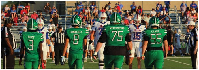 Four people in green football uniforms stand on a field facing away from the camera with a group of people in white uniforms in the background. Descripci n generada con IA