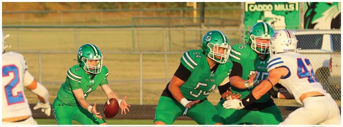 People wearing green and white football uniforms are engaged in a game on a field with a fence and trees in the background. Descripci n generada con IA