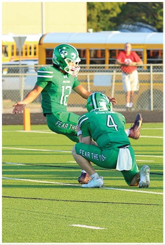 Two people in green football uniforms with helmets are on a field, one holding the ball for the other to kick. Descripci n generada con IA