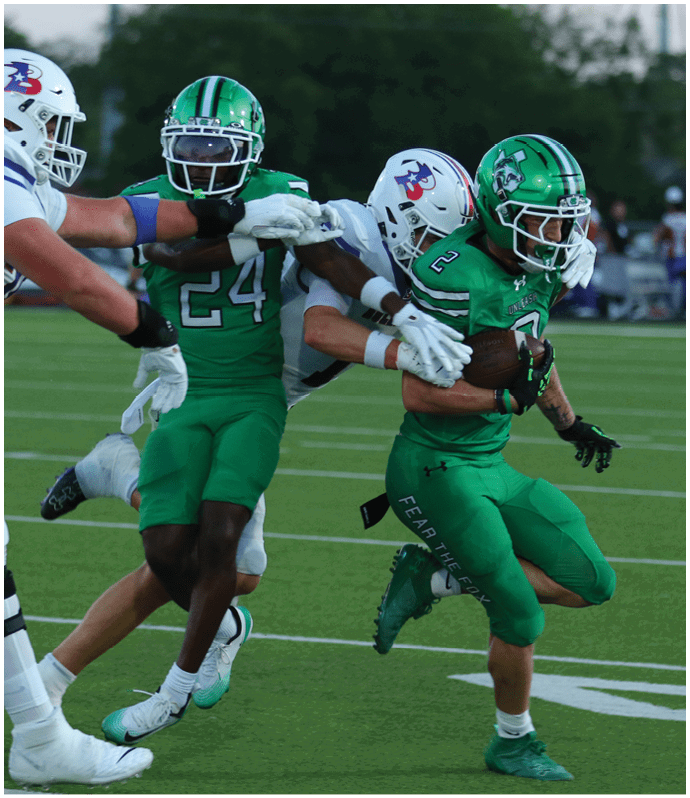 People in football uniforms are actively engaged in a play on a green field with one player in green attempting to move past two players in white. Descripci n generada con IA
