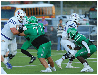 Two football players in green uniforms are tackling or blocking two players in white uniforms during a game on a grassy field with a school bus and spectators in the background. Descripci n generada con IA