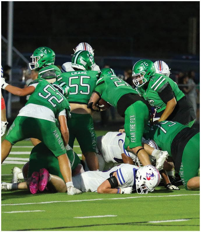 People in green and white football uniforms are engaged in a tackle on a football field during a nighttime game. Descripci n generada con IA