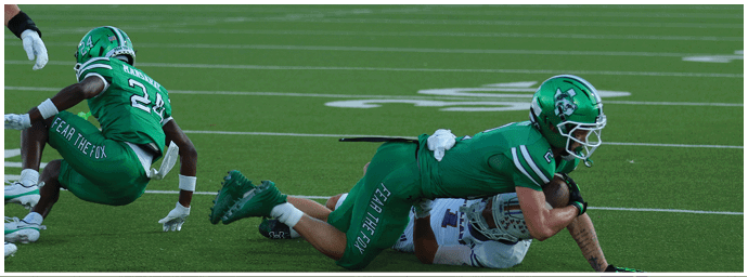 Two football players in green uniforms are on a field with a blurred crowd and band in the background. Descripci n generada con IA