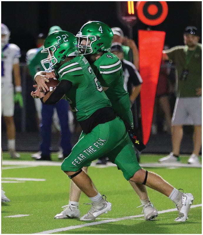 Two people in green football uniforms are engaged in a play on a football field with other players and officials visible in the background. Descripci n generada con IA