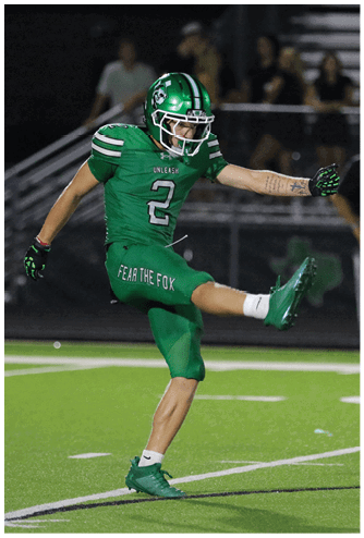 A person in a green football uniform is kicking a football on a field with blurred spectators in the background. Descripci n generada con IA