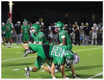 Players in green and white football uniforms are actively engaged in a nighttime game on a well-lit field with spectators in the background. Descripci n generada con IA