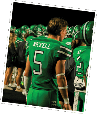 A person wearing a green football uniform with the name Nickell and number 5 stands among a group of people in similar green uniforms and helmets on a sports field at night. Descripci n generada con IA