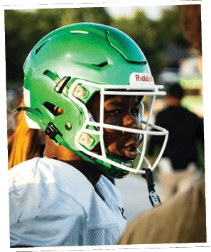 A person wearing a green football helmet and white jersey is looking to the side during an outdoor event. Descripci n generada con IA