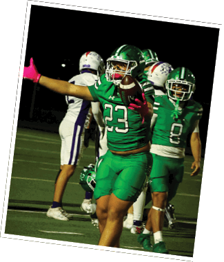 A person in a green football uniform with the number 23 is catching a football while extending one arm, with other players in green and white uniforms visible in the background on a football field at night. Descripci n generada con IA