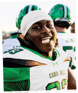 A person wearing a white and green football uniform with the name “Caddo Wolves" is smiling and looking at the camera with other players in green helmets in the background. Descripci n generada con IA
