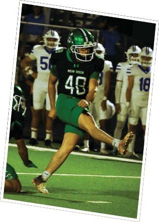 A person in a green and black football uniform is kicking a ball while another person in a similar uniform is crouched nearby, with people in white uniforms standing in the background. Descripci n generada con IA