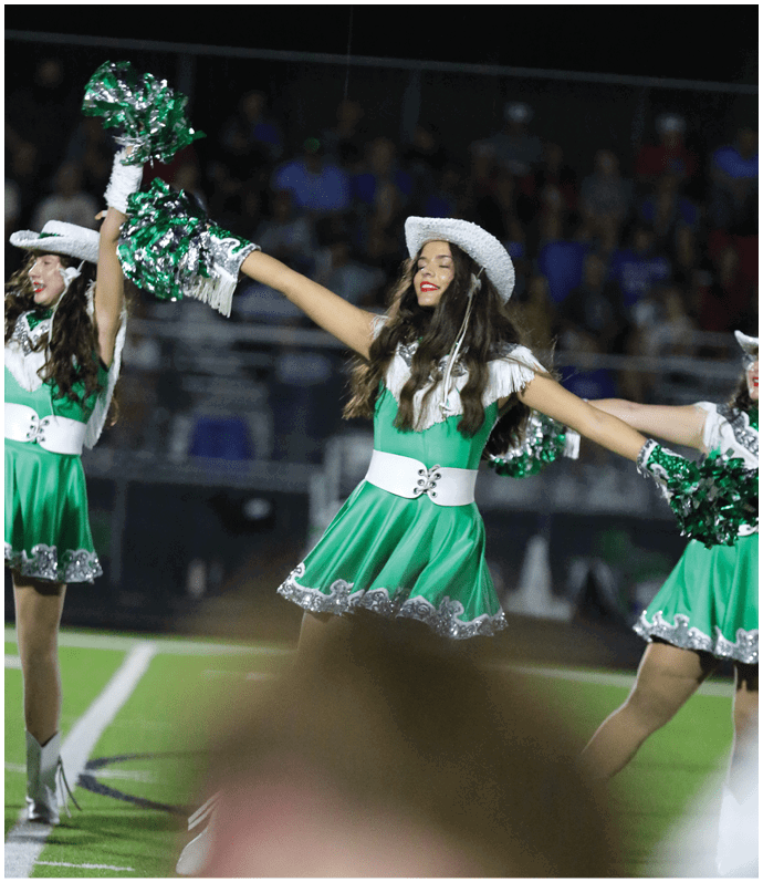 A person wearing a white hat and outfit is smiling and holding up green and silver pom-poms, likely performing at an event with a blurred crowd in the background. Descripci n generada con IA