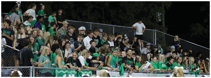 A large group of people wearing green and black clothing are gathered closely together in bleachers at an outdoor event during nighttime. Descripci n generada con IA