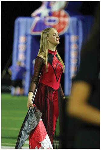 A person with long straight hair wearing a red and black sequined costume is standing with eyes closed and a slight smile, with a blurred background featuring large colorful shapes. Descripci n generada con IA