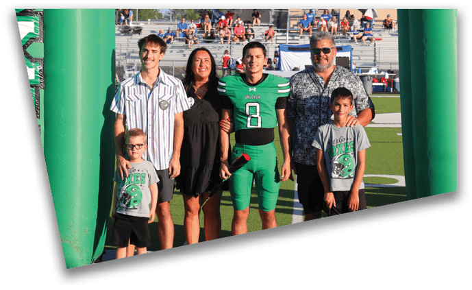 Four adults stand arm in arm on a sports field between two large green inflatable pillars with the word “XES" on them, with spectators visible in the background. Descripci n generada con IA