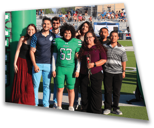 A group of people, including one in a green football uniform, stand together smiling under a large green inflatable arch with the word “FOXES" on it at a sports field. Descripci n generada con IA