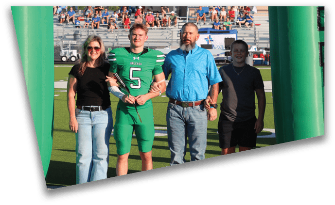 Four adults stand arm in arm on a sports field between two large green inflatable pillars with the word “XES" on them, with spectators visible in the background. Descripci n generada con IA