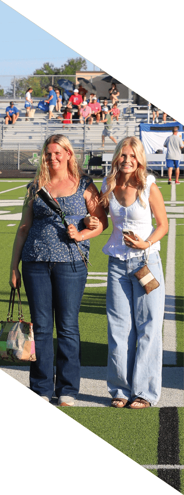 Dos personas adultas sonrientes caminan juntas por un campo deportivo con gradas y espectadores al fondo bajo un cielo despejado. Descripci n generada con IA