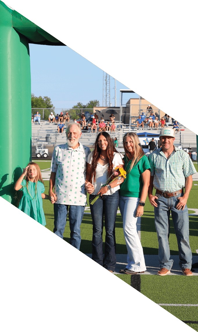 Un grupo multigeneracional de personas sonrientes sostiene flores y se encuentra en un campo deportivo con gradas y espectadores al fondo. Descripci n generada con IA
