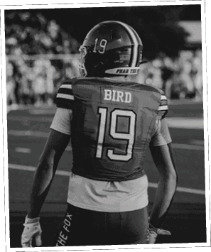 A person wearing a white and green football uniform with the name “Caddo Wolves" is smiling and looking at the camera with other players in green helmets in the background. Descripci n generada con IA