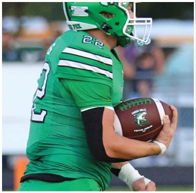 A person in a green football uniform and helmet is running on a field while holding a football. Descripci n generada con IA