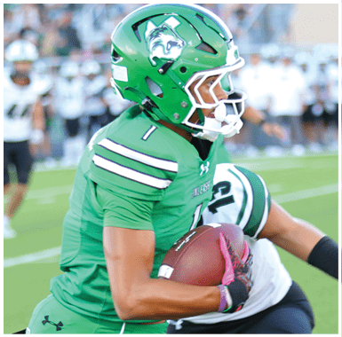 A person in a green football uniform is running with the ball while players in black and white uniforms chase behind on a football field. Descripci n generada con IA